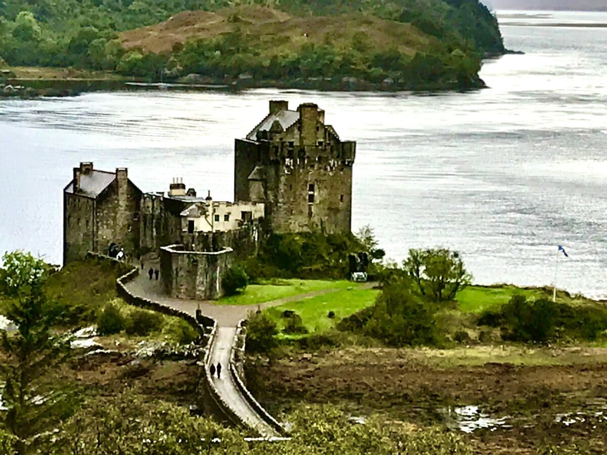 closer view of Eilean Donan Castle from an elevated position, surrounded by the three lochs and mists. A couple walking across the stone bridge towards the entrance
