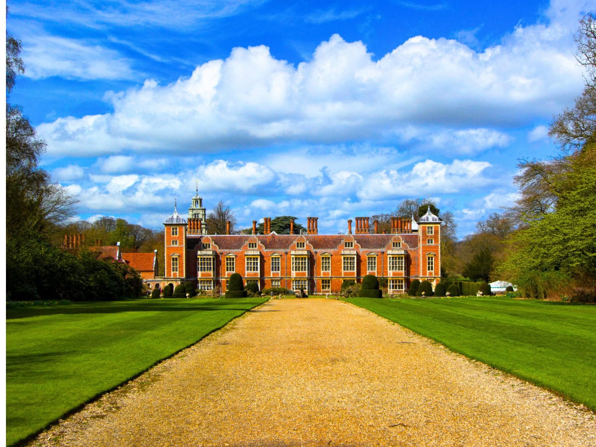 Long path bordered by green lawns leading towards the historic Jacobean façade of Blickling Hall in Norfolk, England.