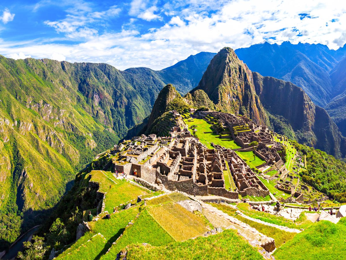 Panoramic view of Machu Picchu, the ancient Incan citadel nestled high in the Andes Mountains of Peru, featuring stone terraces and ruins surrounded by lush green peaks under a partly cloudy sky. ||Guide to Sustainable Travel for Heritage and Culture Explorers + Tips