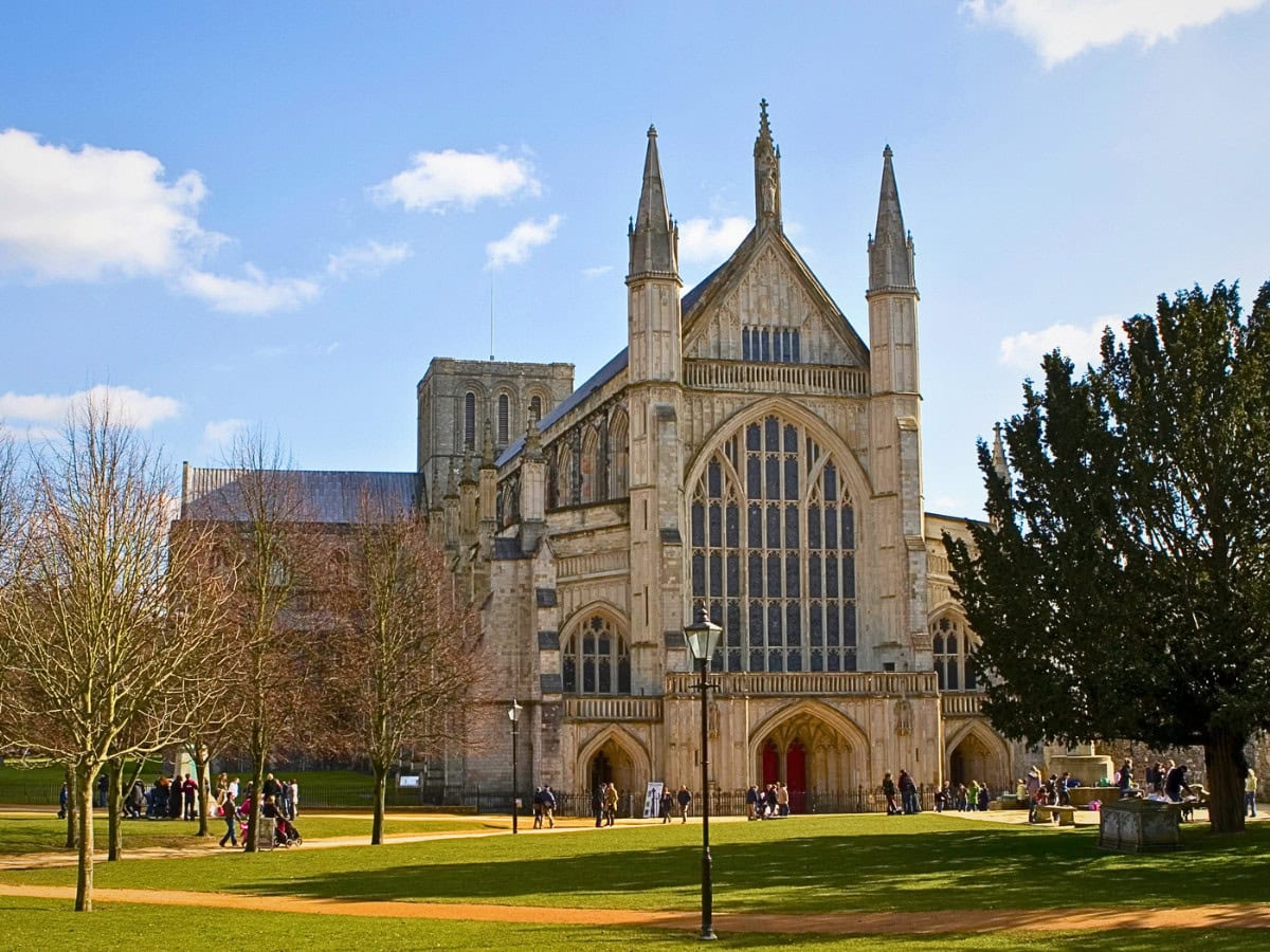 Exterior of Winchester Cathedral with visitors walking around the historic medieval architecture on a sunny day.